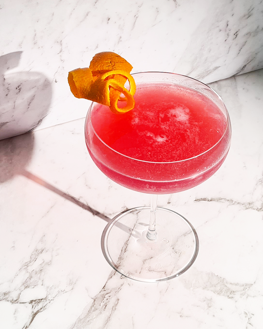 Image of a non-alcoholic cosmopolitan mocktail. It is pink with an orange ring as garnish on the rim of the glass.