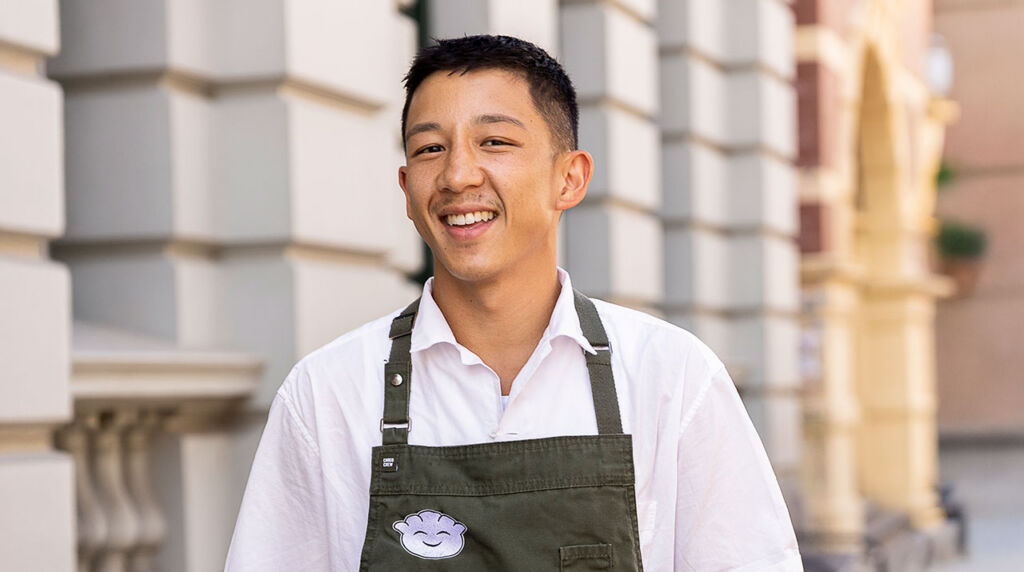 Chef Brendan Pang on the main cooking stage in an apron, smiling while pulling apart dumpling wrapper mix at the Good Food & Wine Show.