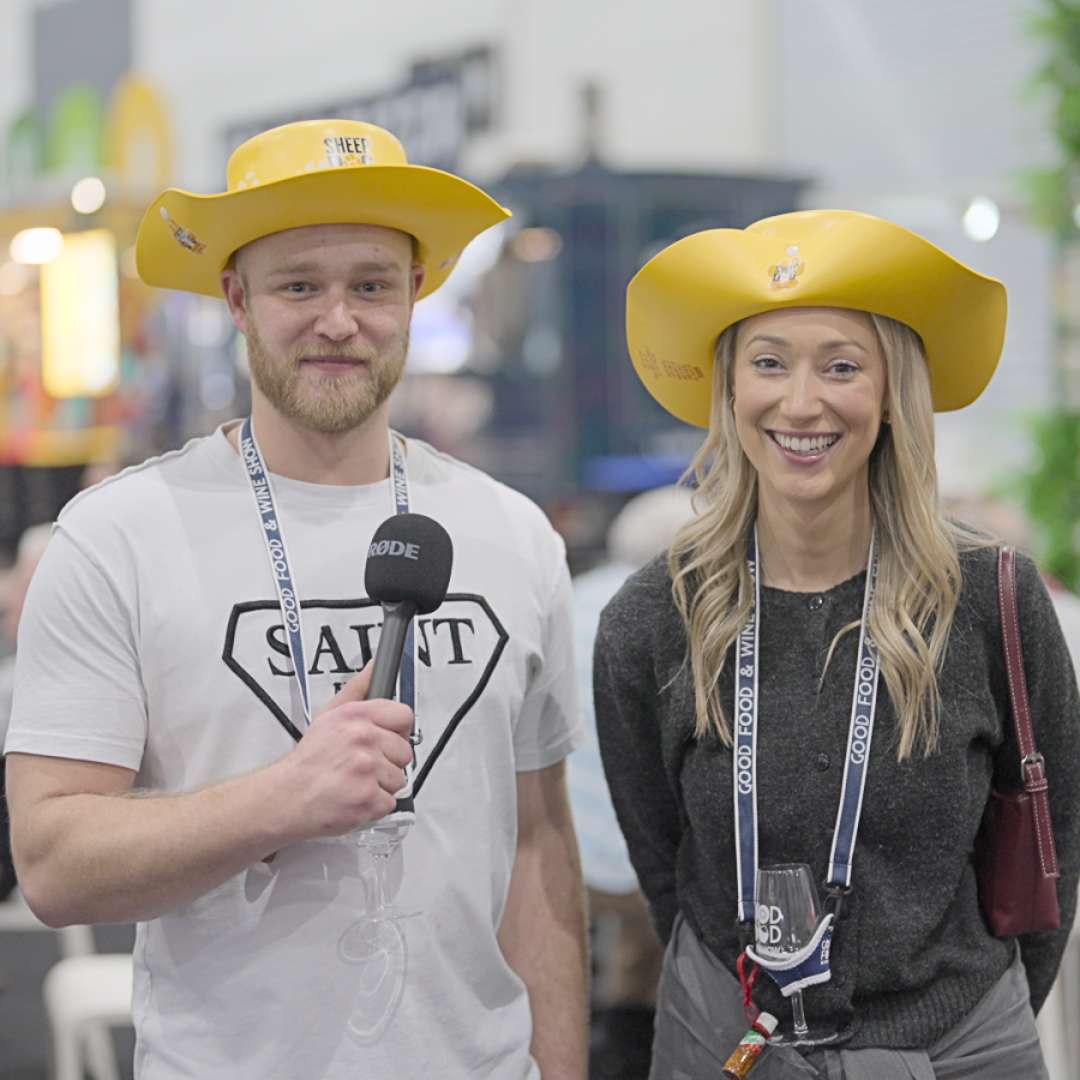 A couple being interviewed at the Good Food & Wine Show, smiling, with the woman wearing a yellow hat.