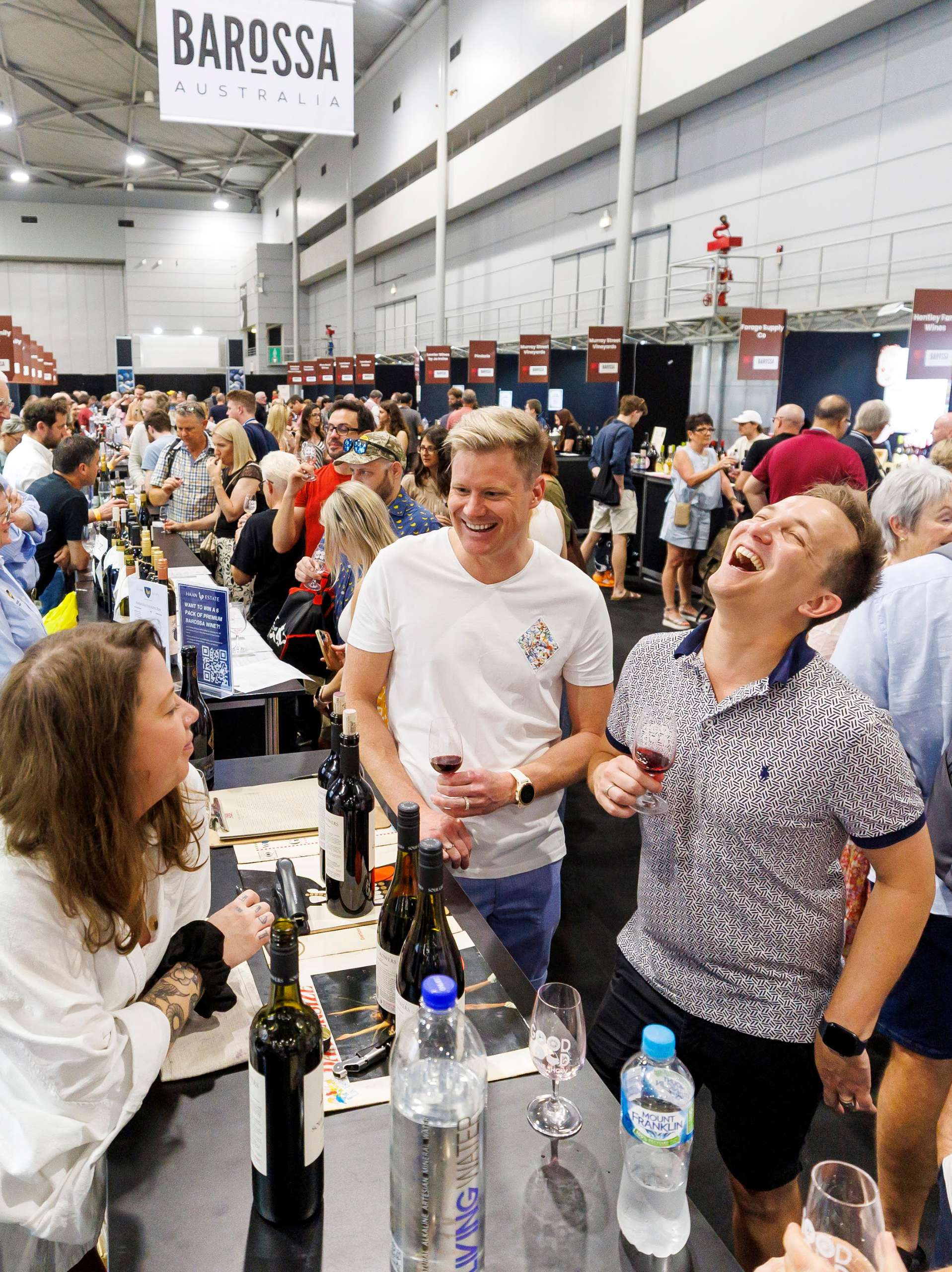 Two men laughing with a winemaker at a Good Food & Wine Show wine pavilion