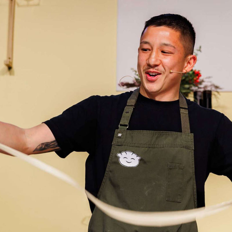 Chef Brendan Pang on the main cooking stage in an apron, smiling while pulling apart dumpling wrapper mix at the Good Food & Wine Show.