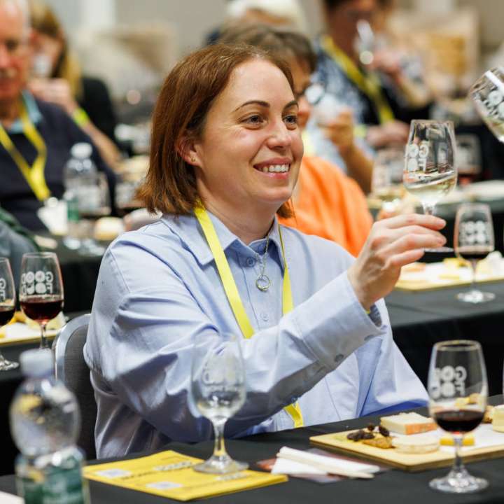 Woman raising a champagne glass during a Cheese, Champagne and Caviar Masterclass at the Good Food & Wine Show, seated at a tasting table with cheese and wine.