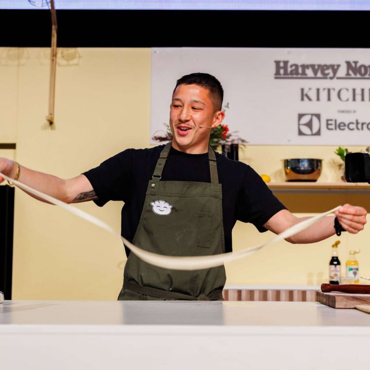 Chef Brendan Pang on the main cooking stage in an apron, smiling while pulling apart dumpling wrapper mix at the Good Food & Wine Show.